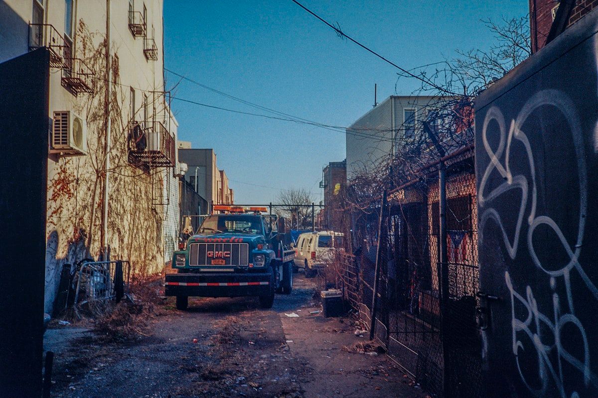 A roll-off dumpster parked in a driveway beside piles of mixed renovation debris such as broken drywall, wood scraps, old flooring, and a stack of roofing shingles, with a tape measure and a calendar nearby to suggest size, weight, and rental period considerations