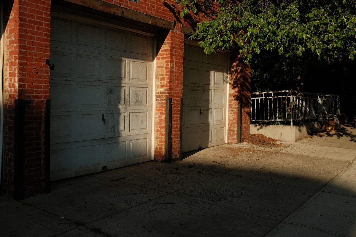 A residential driveway with a pile of removed kitchen cabinets, a stack of flooring boards, a few old furniture pieces, and a roll-off dumpster parked beside the garage while a tape measure lies on the driveway near the debris.