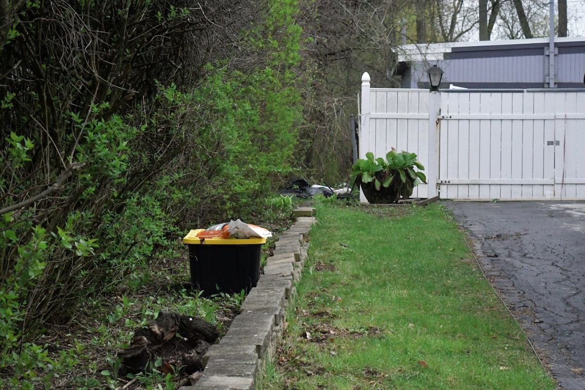 A driveway scene with four roll-off dumpsters lined up in different sizes, each partially filled with matching debris: one with bathroom demolition scraps and broken tile, one with stacked garage clutter and flooring pieces, one with kitchen cabinets and drywall, and one with bulky furniture and loose boxes, showing the visual difference between 10-yard, 15-yard, 20-yard, and 30-yard containers.