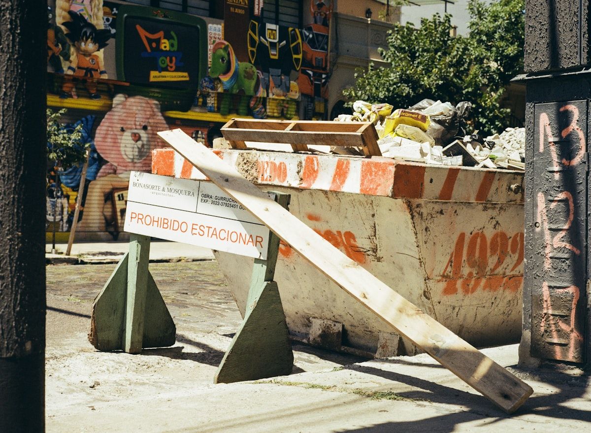 Two dumpsters at a construction site, one filled with bulky household junk like a couch and chairs and the other loaded with heavy debris like bricks, dirt, and shingles, with a narrow driveway and delivery truck in the background to show how material type and access affect cost
